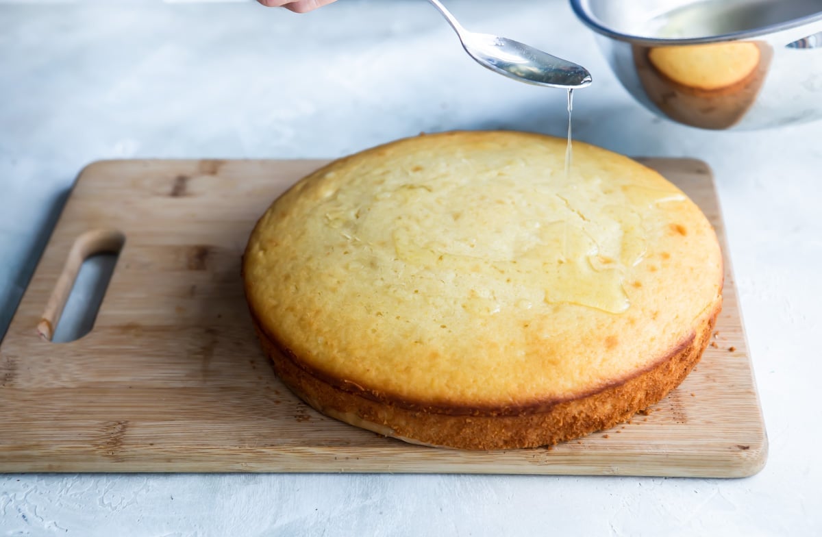 A clear liquid being drizzled over a lemon cake on a wooden cutting board.