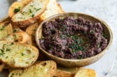 Homemade olive tapenade in a bowl surrounded by toasted bread.