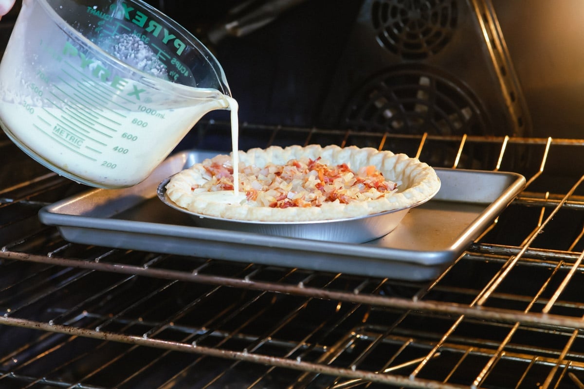 Someone pouring white liquid into quiche Lorraine that is in the oven.
