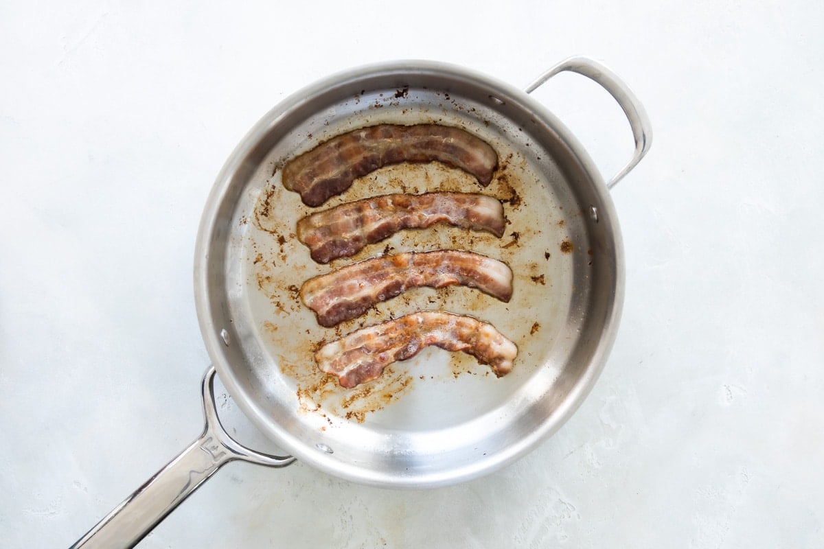 Four slices of bacon being cooked in a silver skillet.