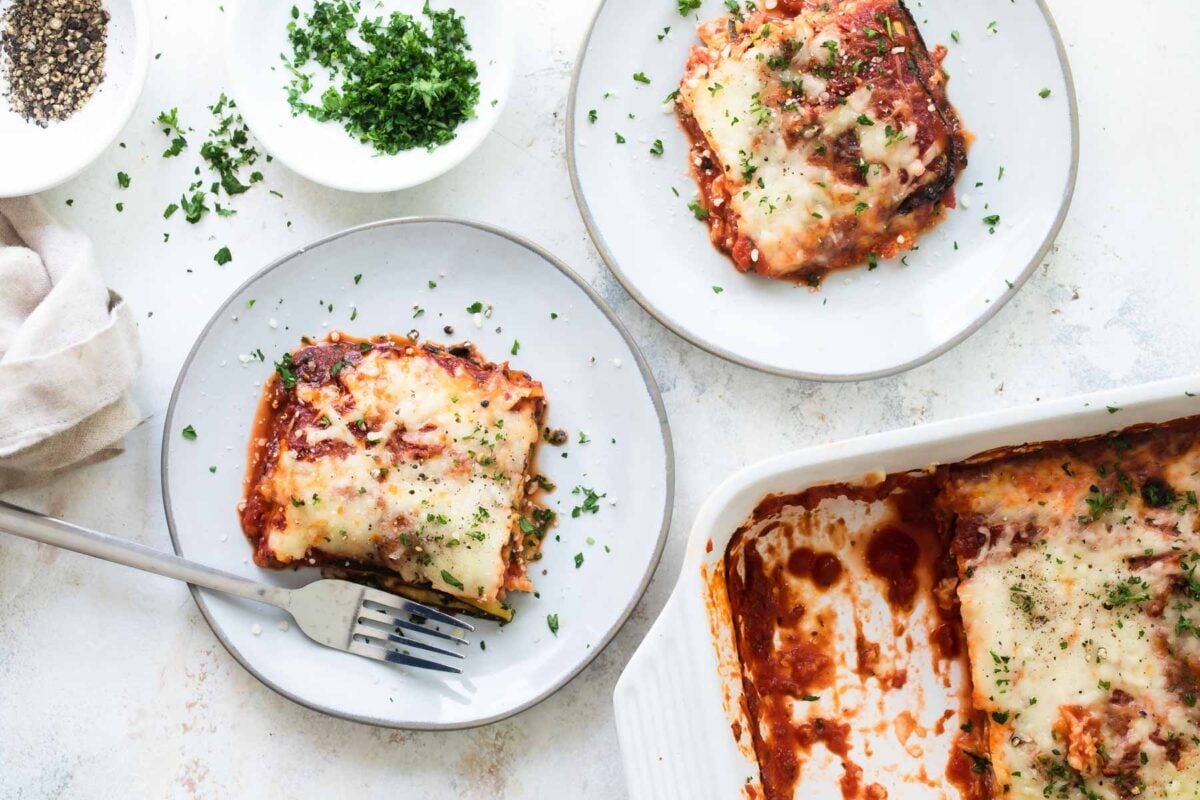 Slices of Zucchini Parmesan on two plates next to a baking dish of Zucchini Parmesan.