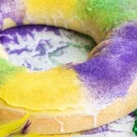 A decorated king cake on a white counter surrounded by Mardi Gras decorations.