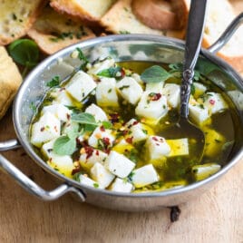 Marinated feta in a silver bowl on a wood board with slices of bread.