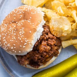 Plates of Sloppy Joes on plates with chips and pickles nearby.