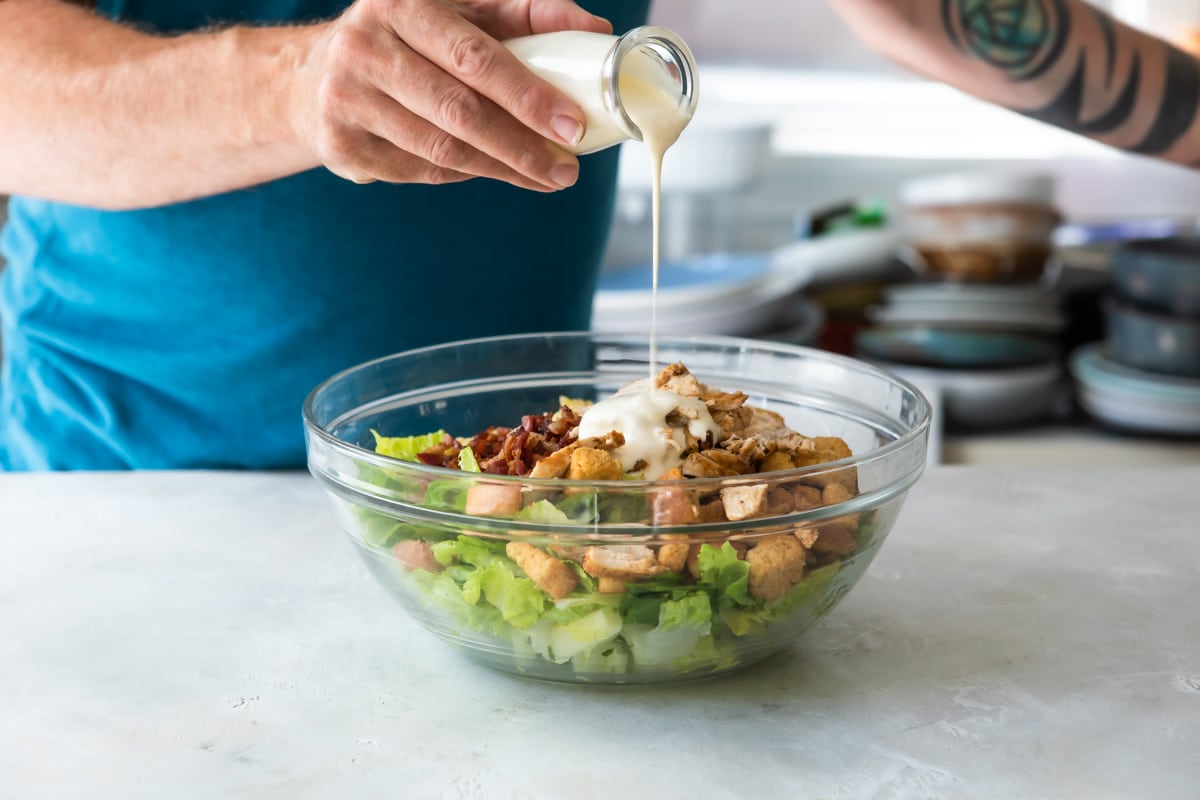 Someone pouring Caesar dressing into a bowl with chicken Caesarsalad.