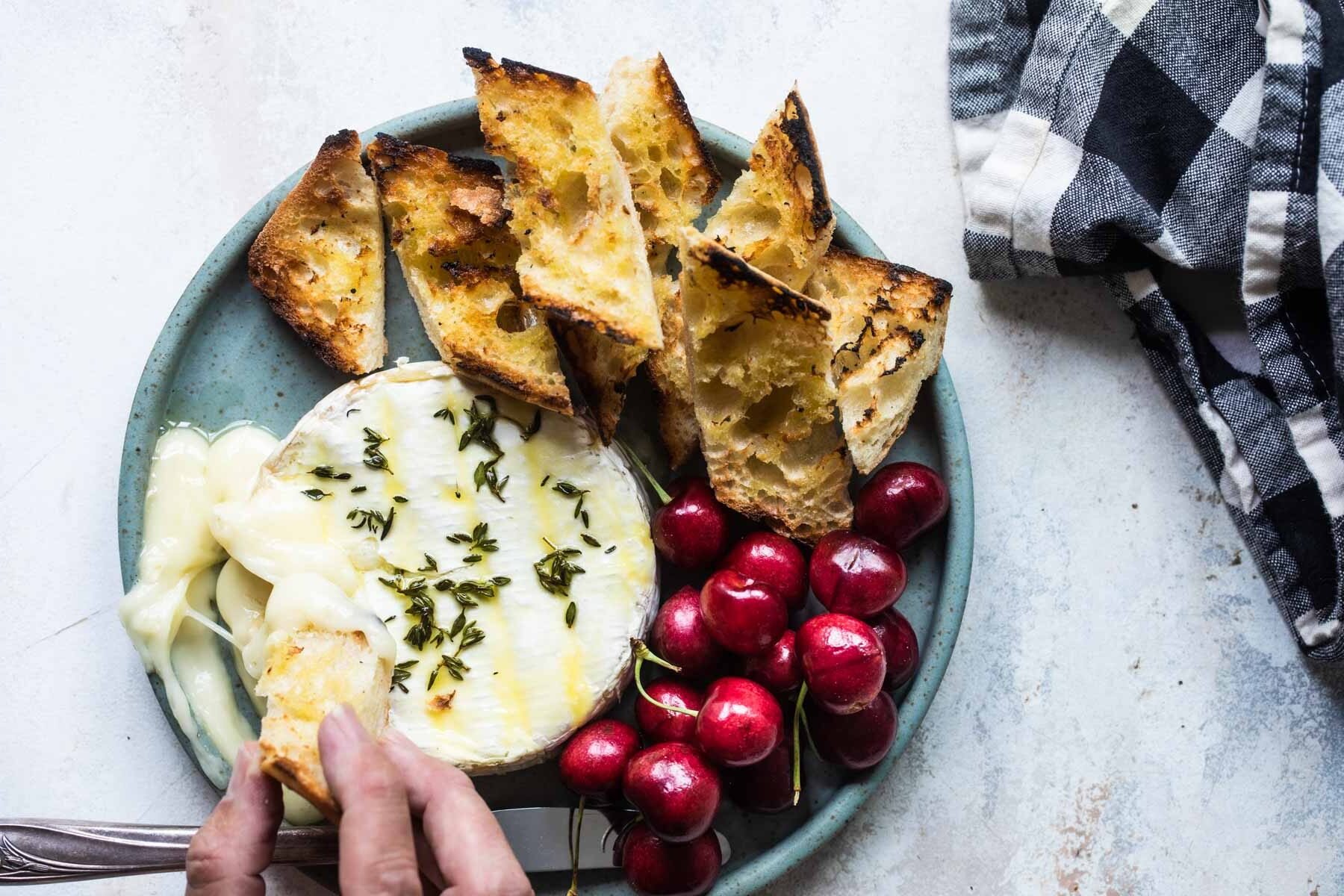 Baked brie on a blue plate with cherries and toasted bread pieces.