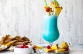 A Blue Hawaiian cocktail surrounded by pineapple and maraschino cherries.
