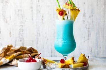 A Blue Hawaiian cocktail surrounded by pineapple and maraschino cherries.