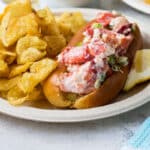 White plates with lobster rolls, potato chips, and lemon slices piled on top.
