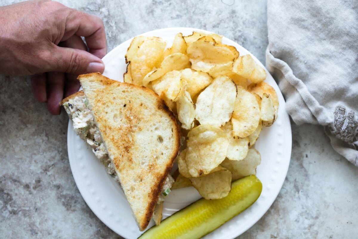 A tuna salad sandwich on a plate next to chips and a pickle spear.