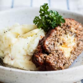 A slice of cheesy meatloaf with mashed potatoes in a low gray bowl.