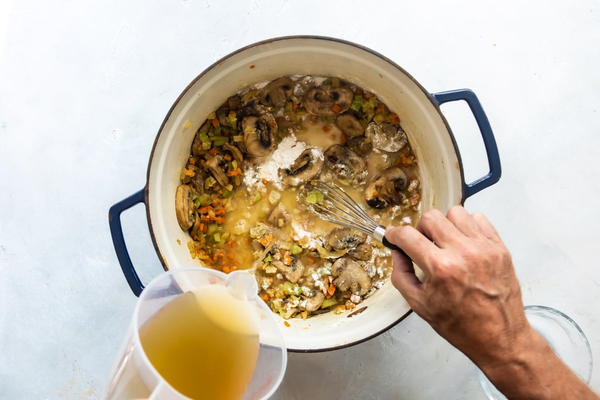Someone stirring chicken broth into the vegetable mixture to make chicken wild rice soup.
