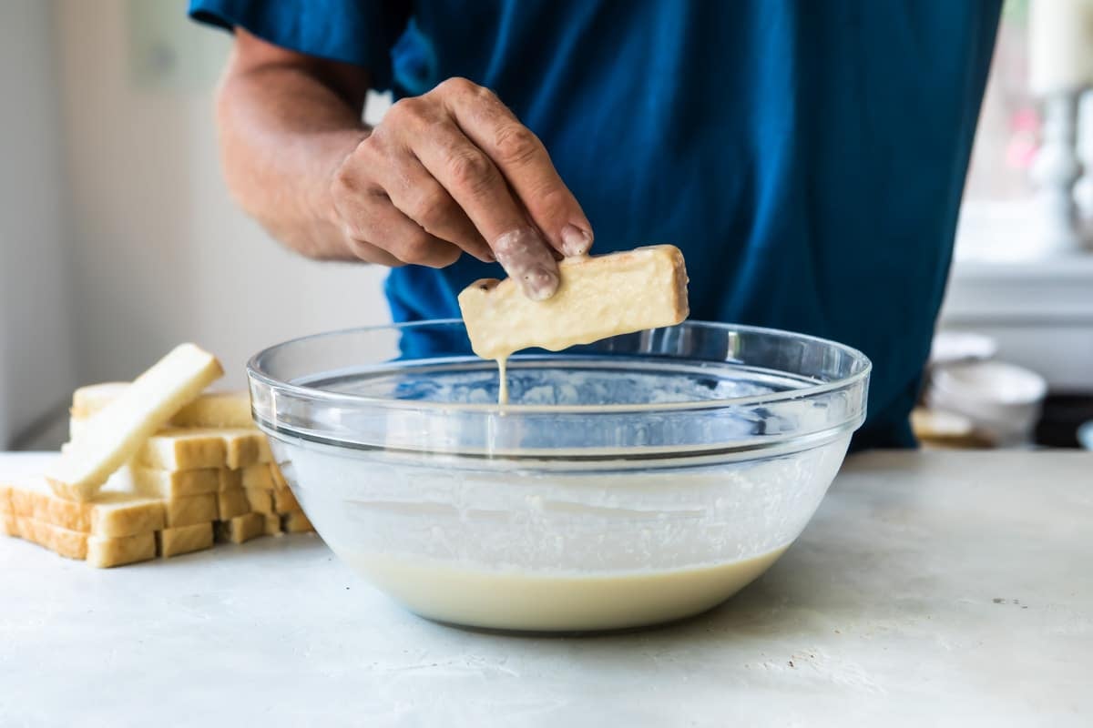 Someone dipping bread slices in a bowl of batter to make French toast sticks.