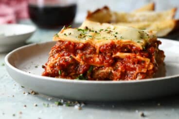 A slice of lasagna on a plate. Wine and garlic bread in the background.