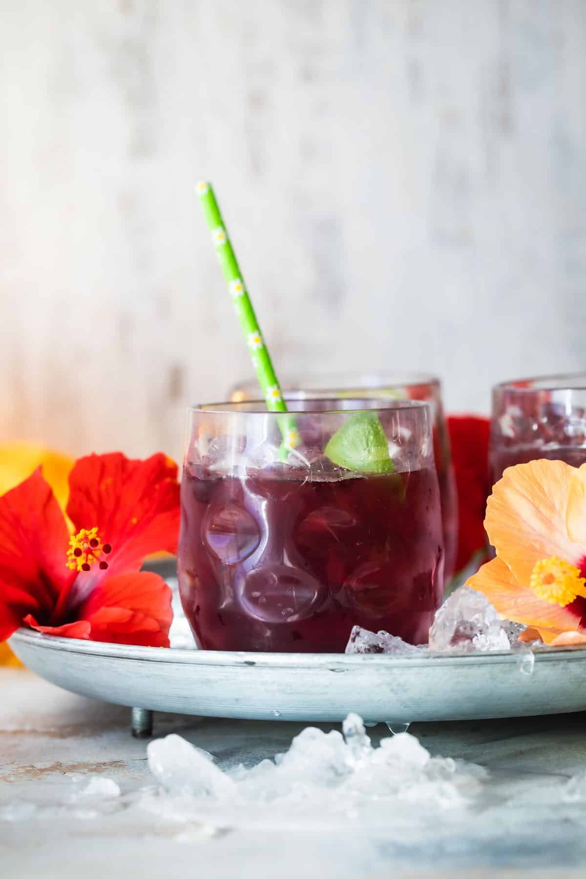 Glasses of Agua de Jamaica on a tray with hibiscus flowers and straws.