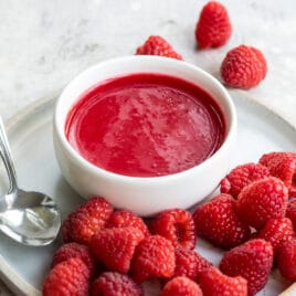 Raspberry coulis in a white dish on a white plate with raspberries.