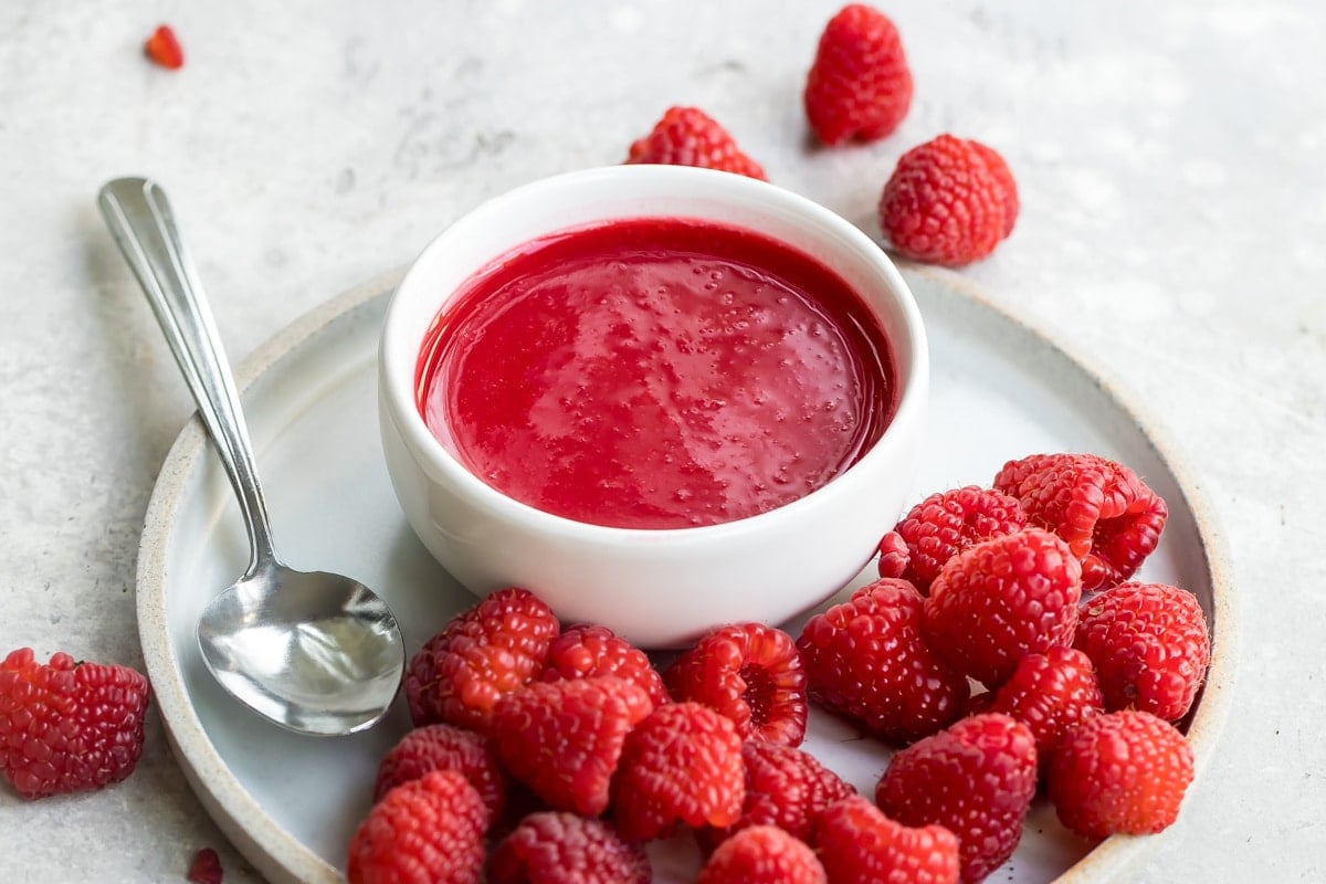Raspberry coulis in a white dish on a white plate with raspberries.