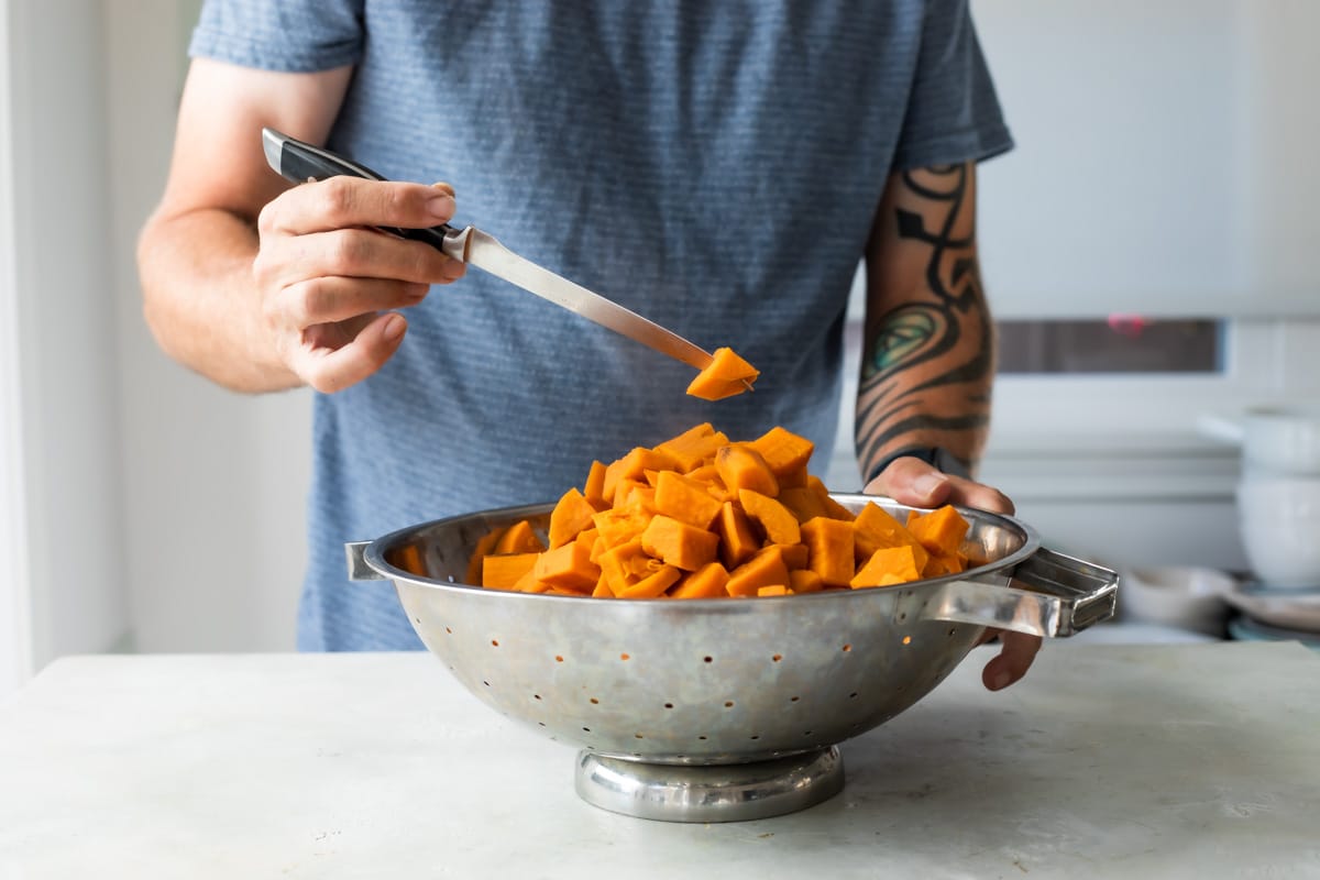 A potato cube getting poked with a knife over a colander of sweet potato cubes.