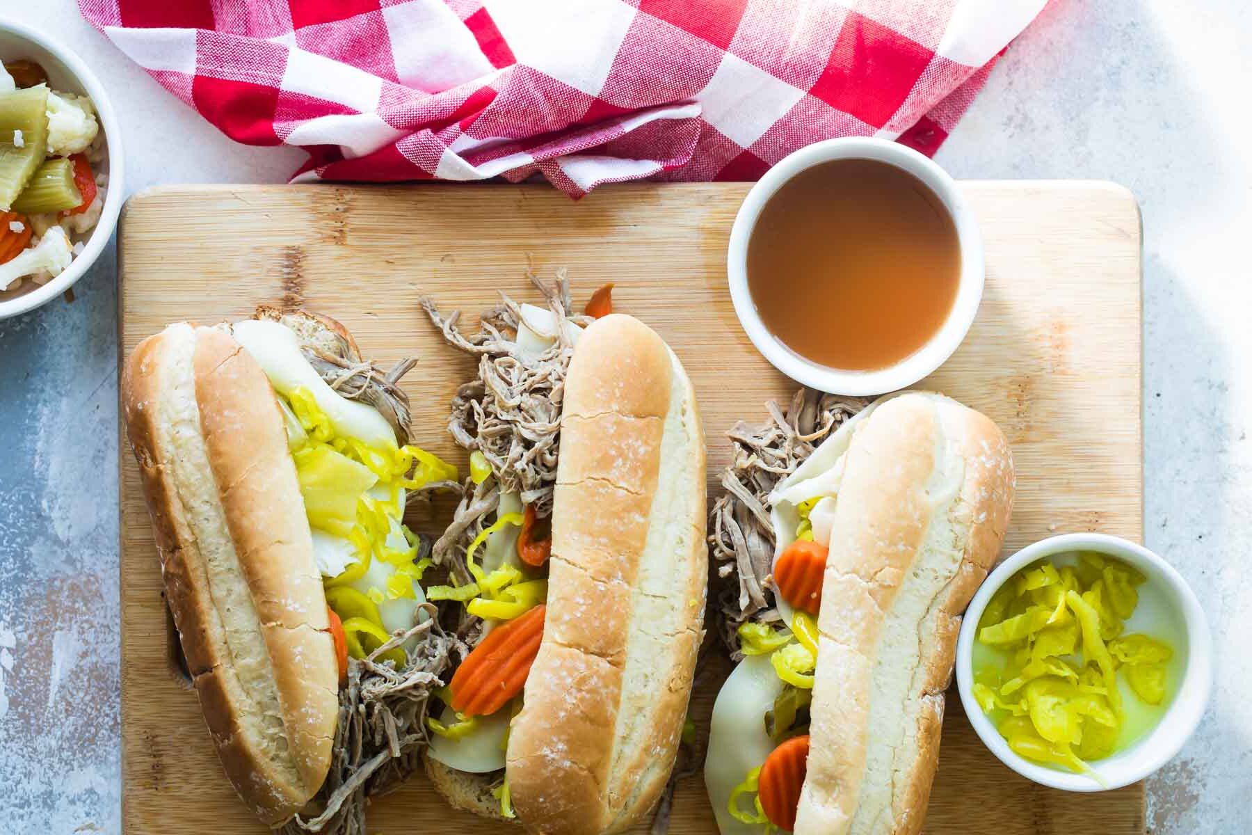 A cutting board with several Italian Beef sandwiches and a bowl of au jus nearby.