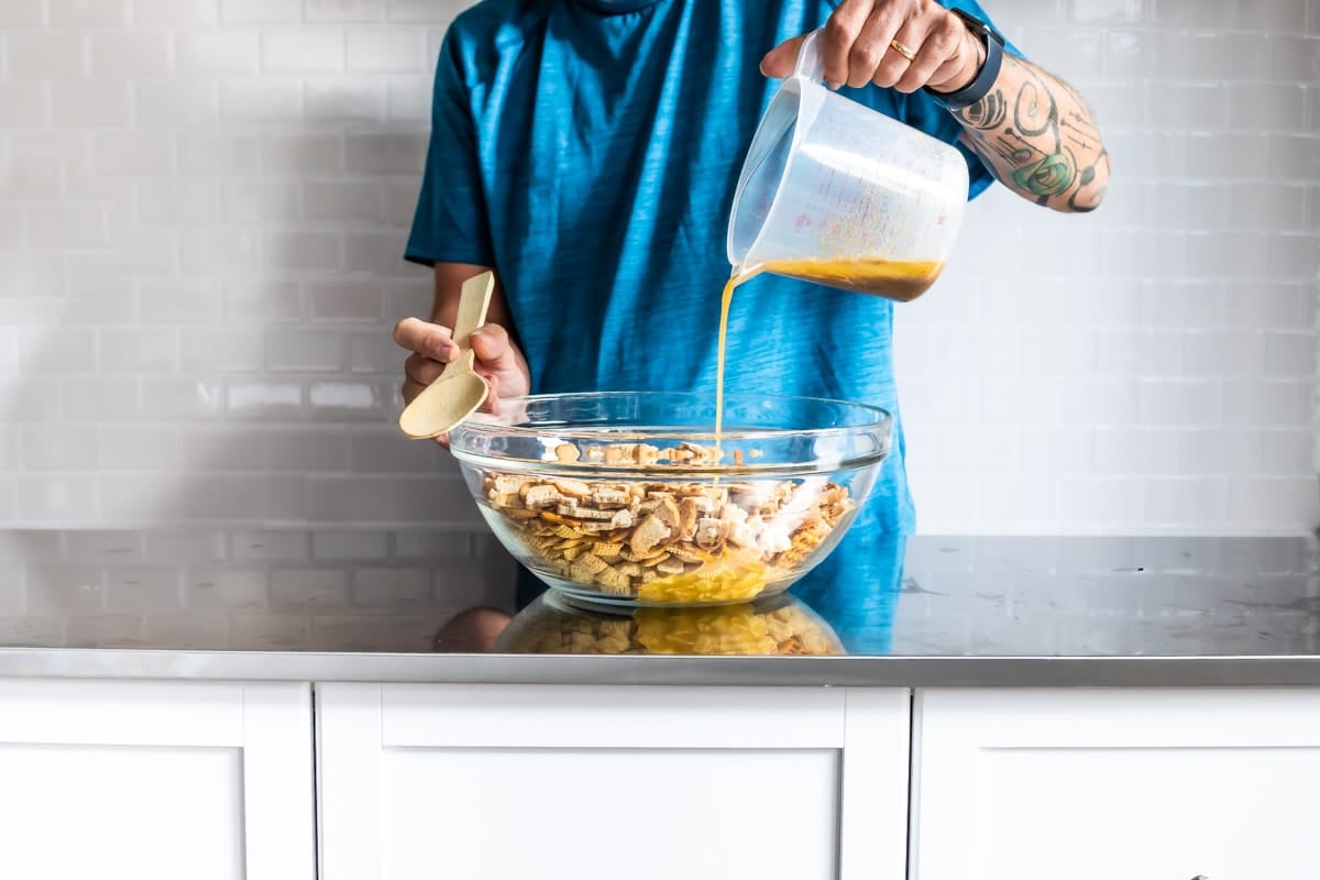 Someone pouring a wet mixture into a bowl of dry chex mix.