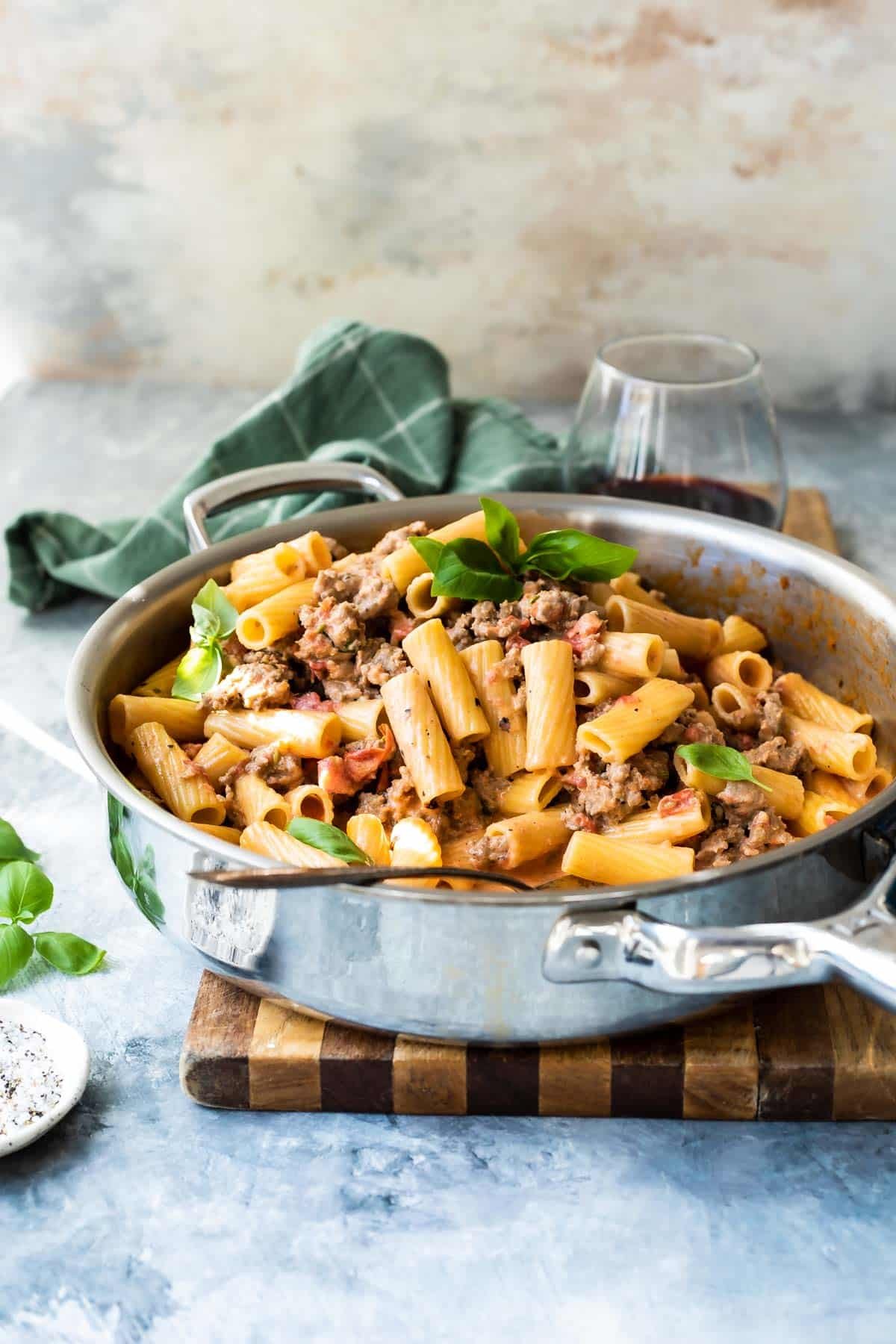 A large skillet filled with Rigatoni with Sausage with a green napkin and wine next to it.