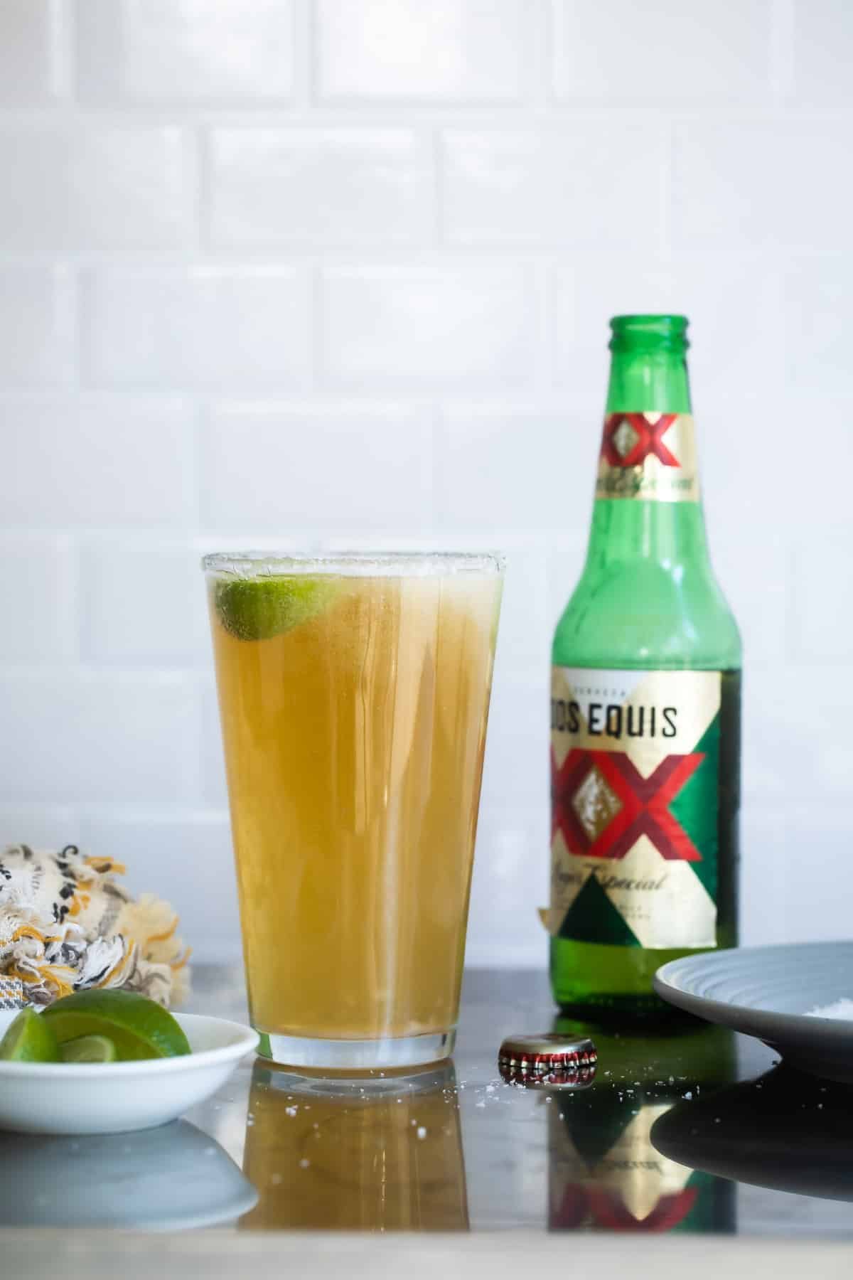 A Chelada next to a beer bottle and a small bowl of limes.