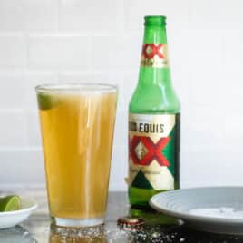 A Chelada next to a beer bottle and a small bowl of limes.