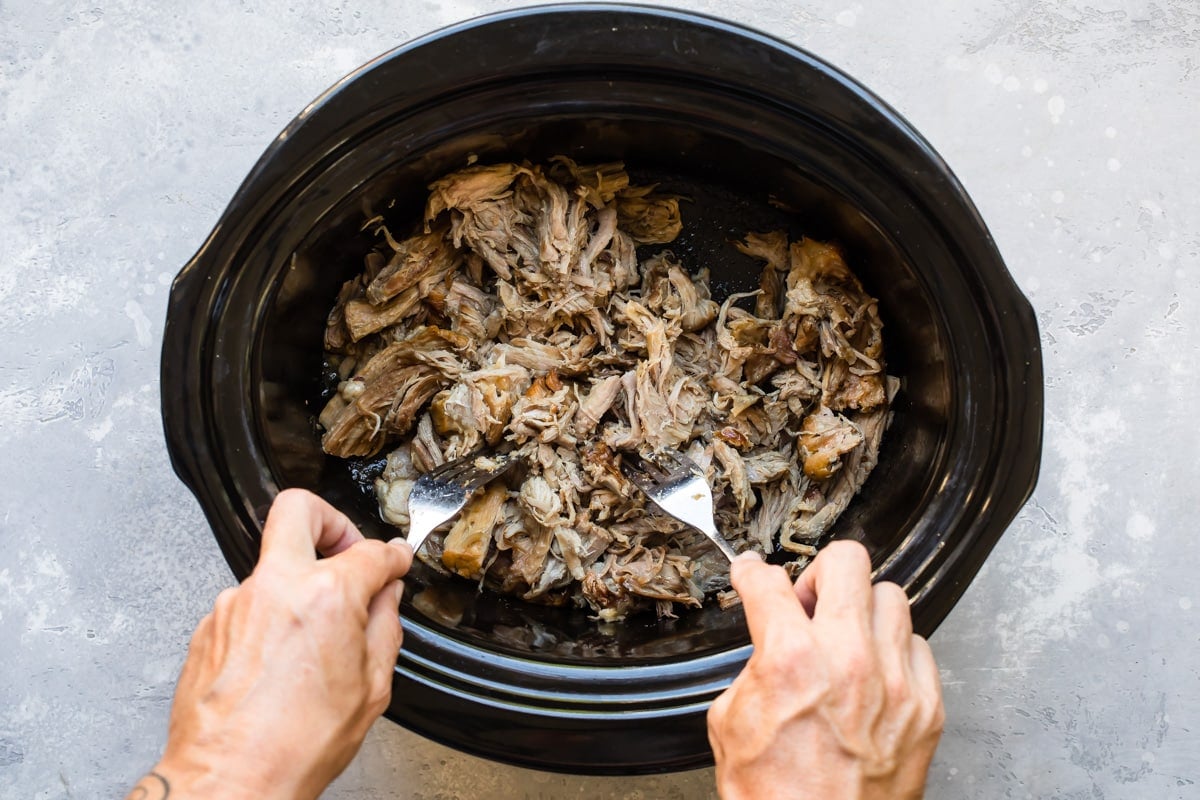 Someone pulling pork in a black slow cooker.