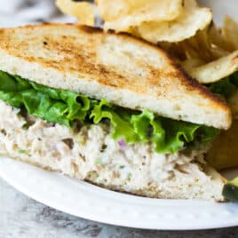 A tuna salad sandwich on a plate with a pickle spear and potato chips.