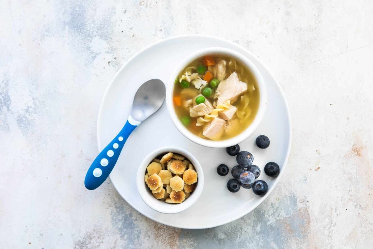 A toddler lunch of chicken noodle soup with a side of blueberries and oyster crackers.