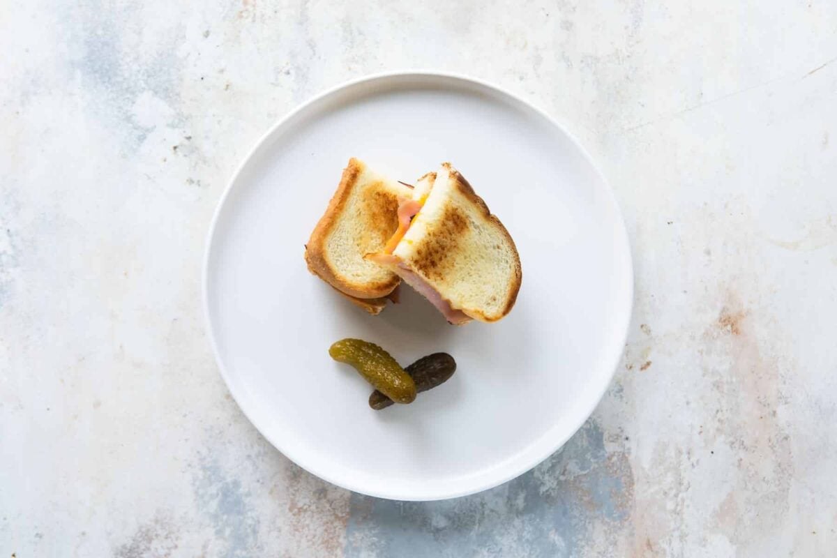 A toddler lunch of a grilled ham and cheese sandwich with a side of pickles.