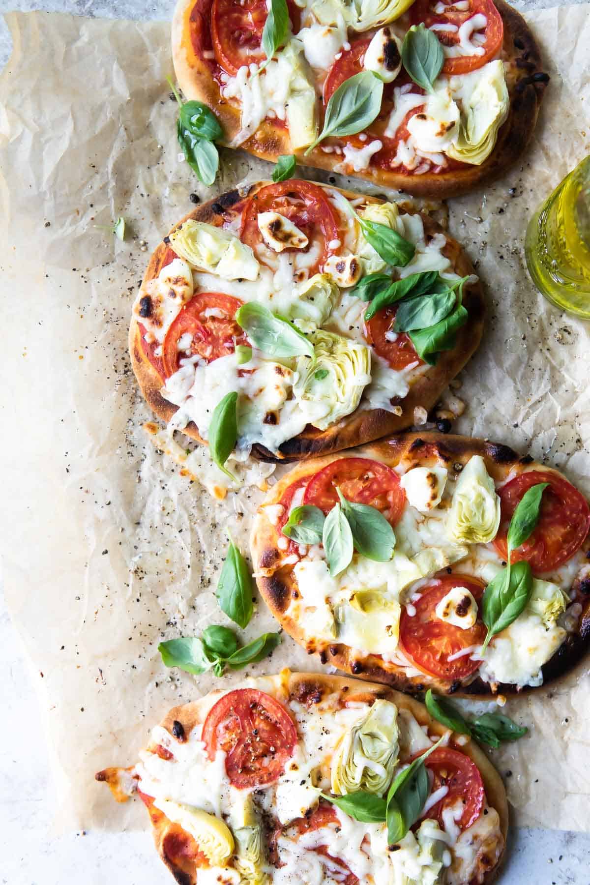 Flatbread pizzas topped with artichokes, tomatoes, cream cheese, and basil leaves on a board.