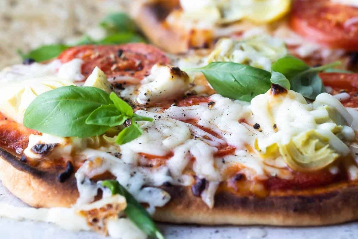 Flatbread pizzas topped with artichokes, tomatoes, cream cheese, and basil leaves on a board.