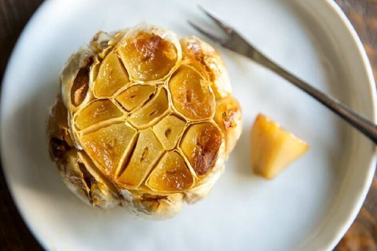 A bulb of roasted garlic on a white plate with an appetizer fork nearby.