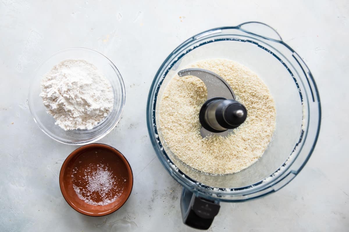 A food processor with some dry ingredients next to a bowl of flour and a bowl of salt.