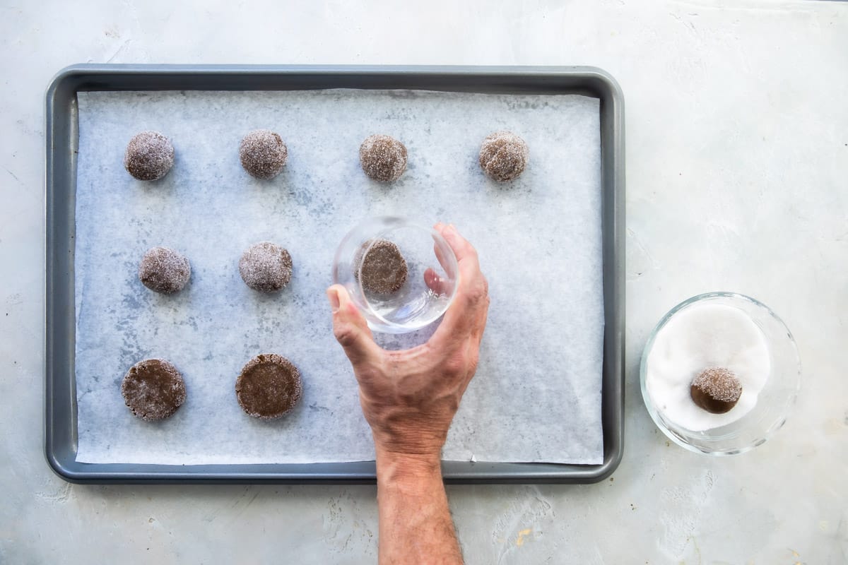Someone flattening molasses cookies with a clear glass on a baking sheet..