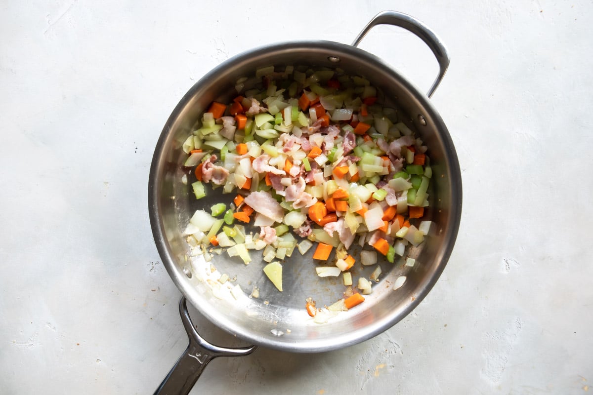 Vegetables and bacon being cooked in a silver skillet to make pasta and bean soup.