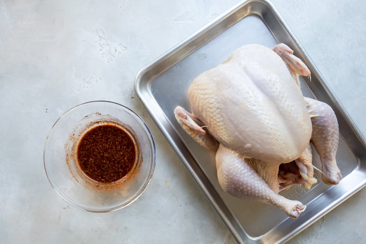A raw chicken on a baking sheet next to a small bowl of seasoning.