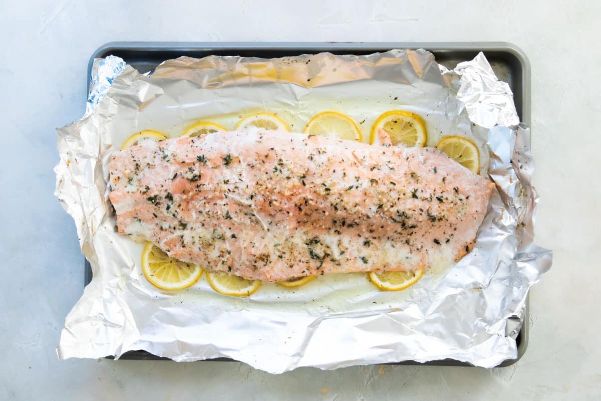 A large side of cooked salmon with spices on top sitting on a layer of lemon slices on top of foil on a baking sheet.
