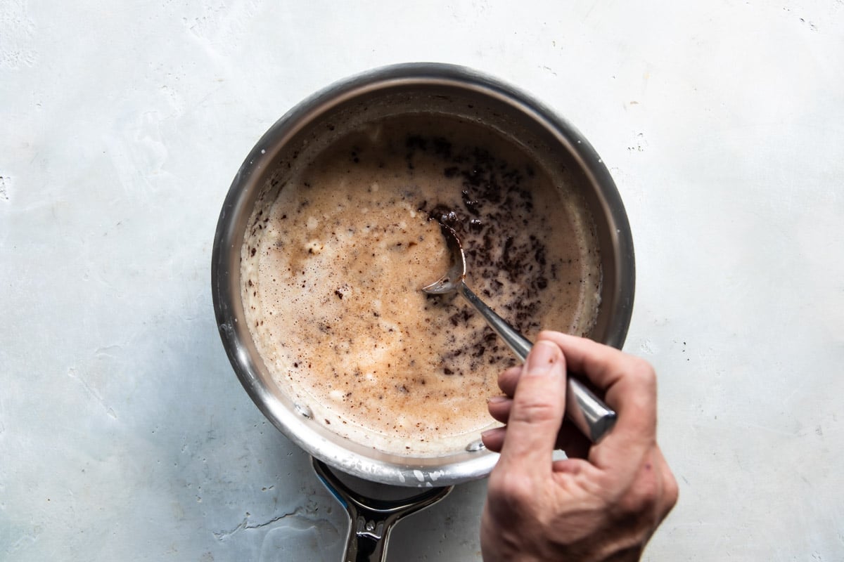 Someone stirring ingredients for chocolate syrup in a metal saucepan.