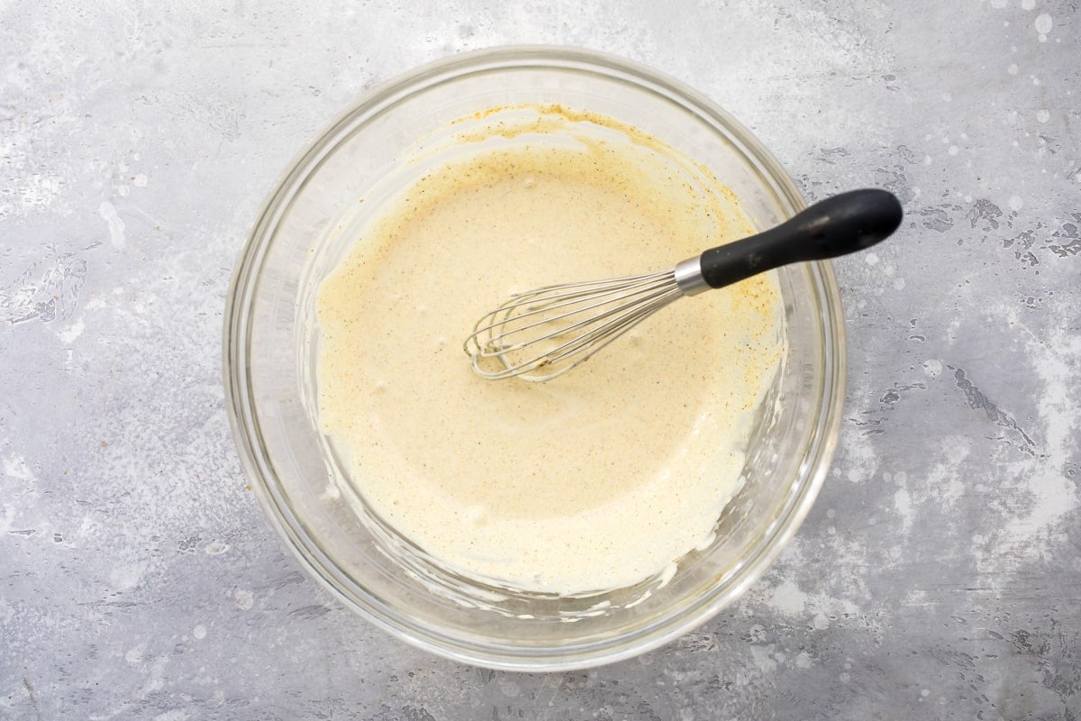 The sauce for curried chicken salad in a clear glass bowl with a whisk in it.