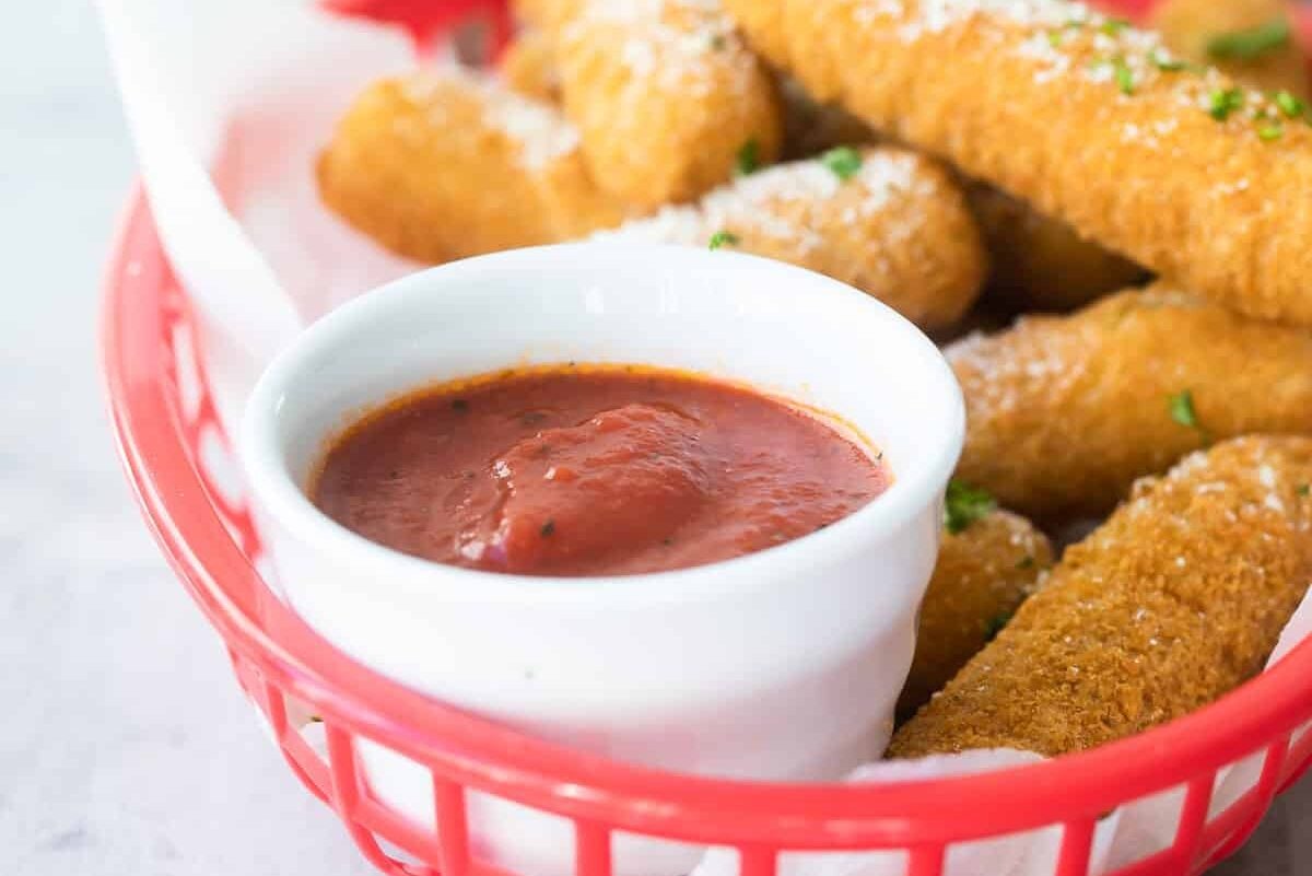 A bowl of marinara in a red plastic basket of mozzarella sticks.