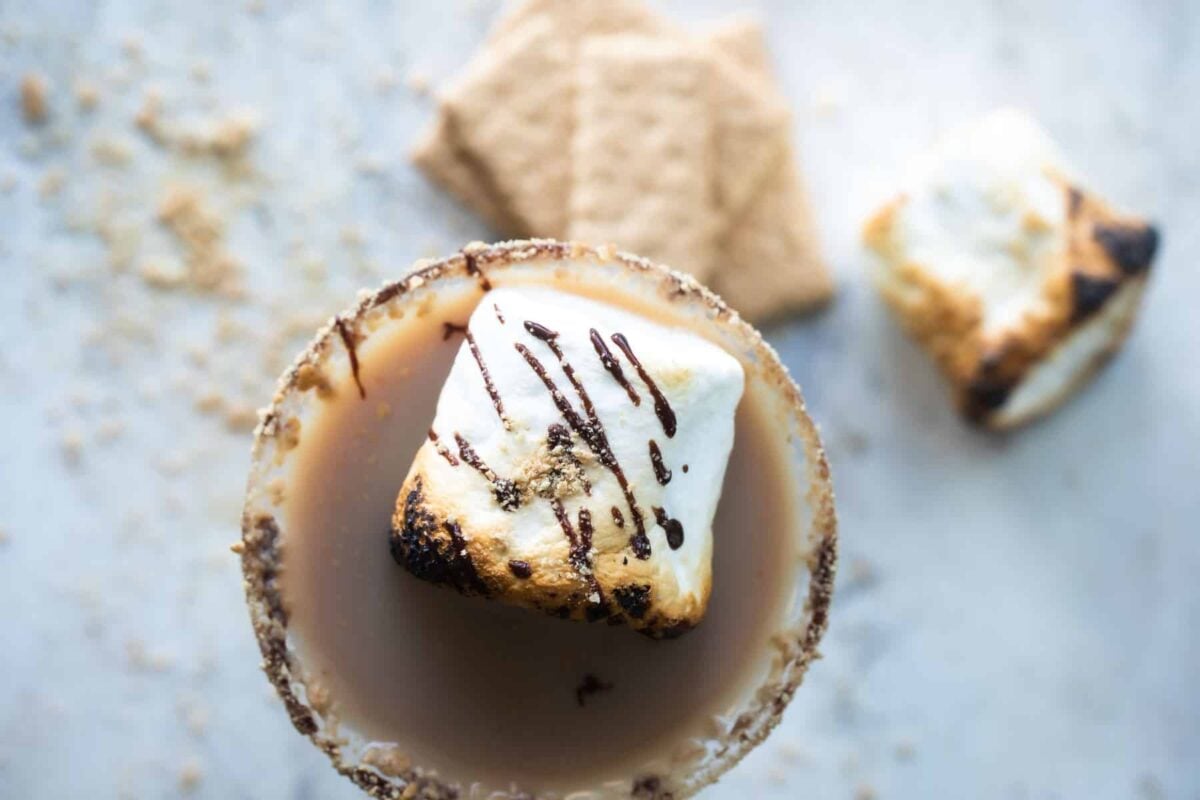 A S'mores Martini in a martini glass next to some graham crackers and a toasted marshmallow.