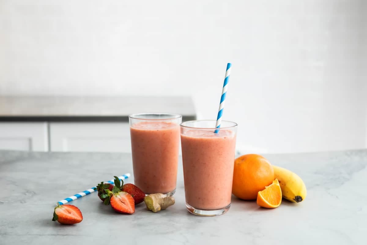 2 glasses with straws of Strawberry Citrus Ginger Smoothie next to fresh fruit.