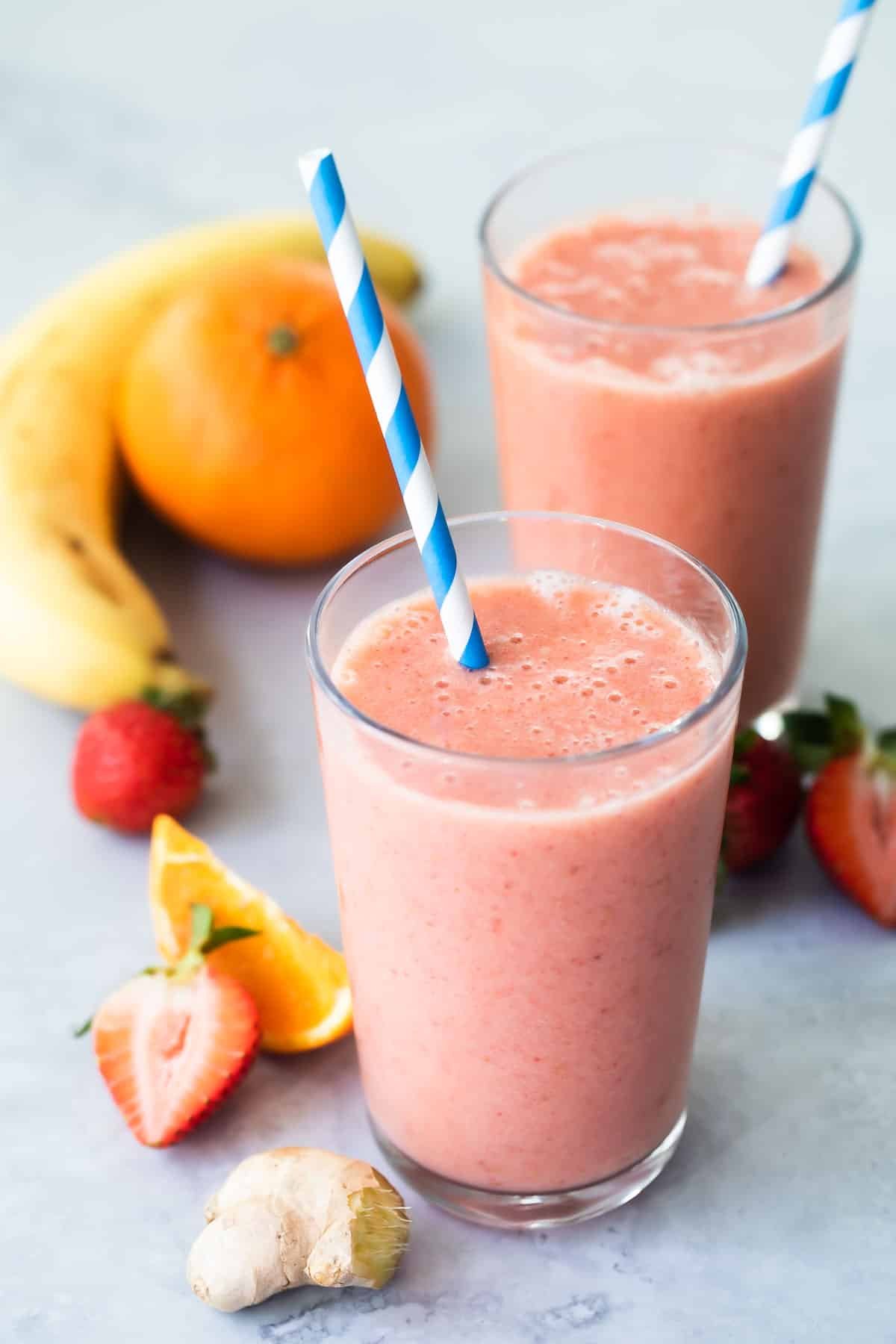2 glasses with straws of Strawberry Citrus Ginger Smoothie next to fresh fruit.