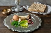A silver Seder Plate with the seder components and a cup of wine and Matzah in the background.