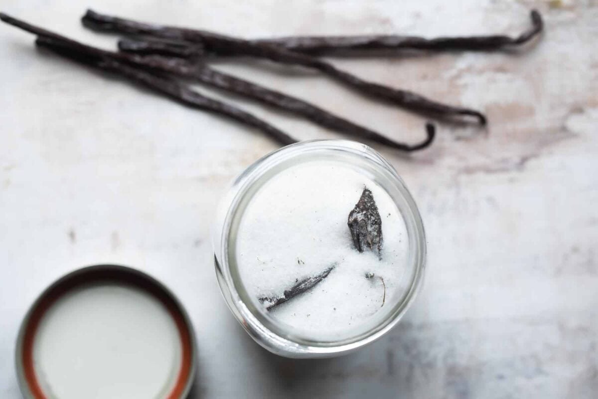 Vanilla sugar in a glass jar next to four vanilla beans.