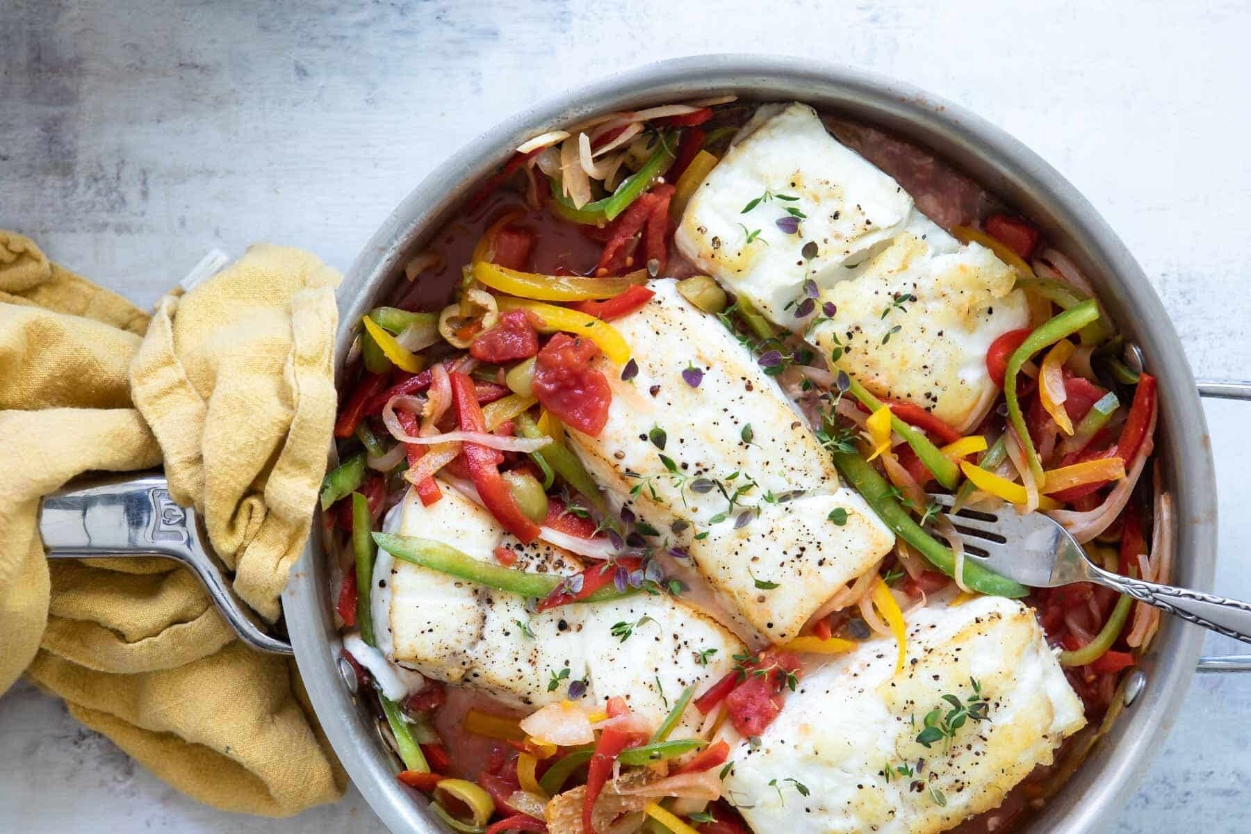 A skillet full of seared halibut, sliced bell peppers and green olives.