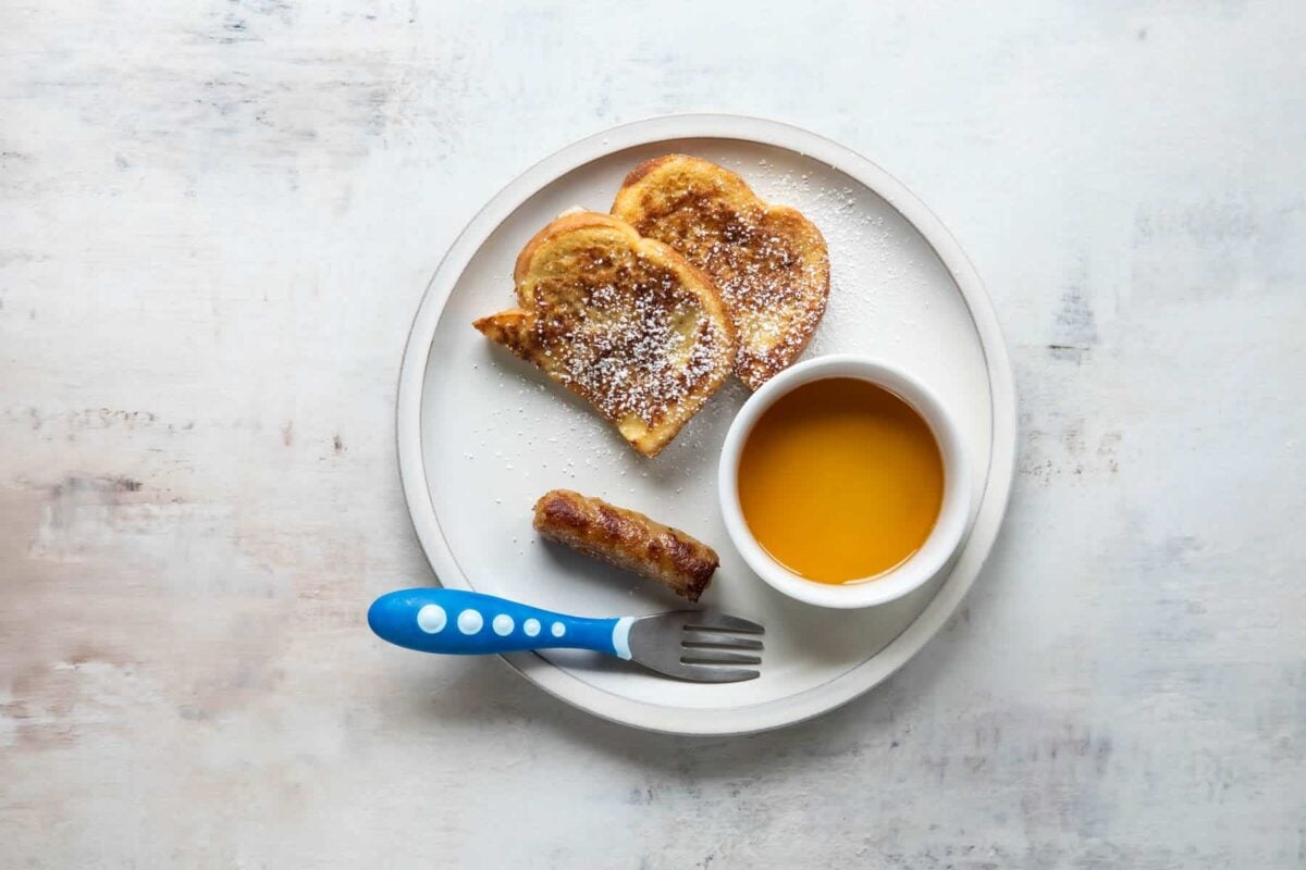 A plate with French toast, syrup, and a piece of sausage on it.