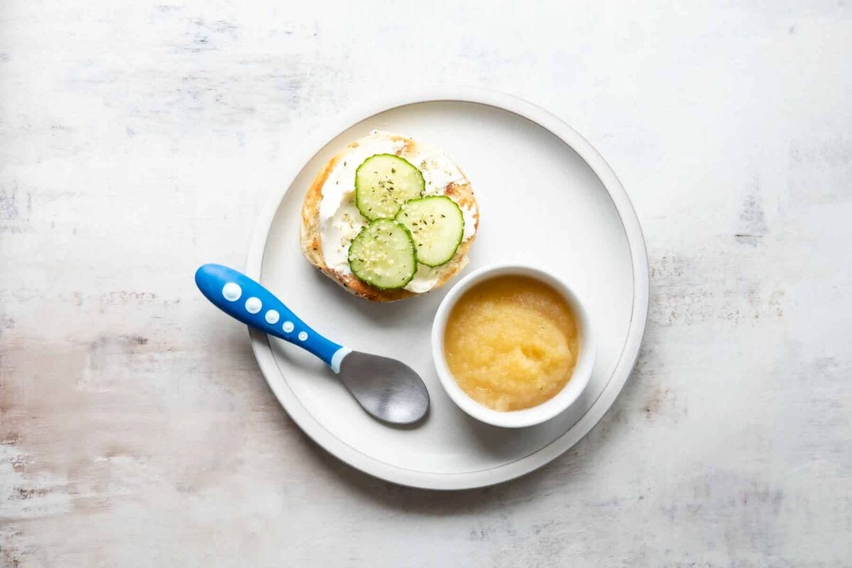 A plate with half of a bagel with cream cheese and cucumbers next to a small bowl of applesauce.
